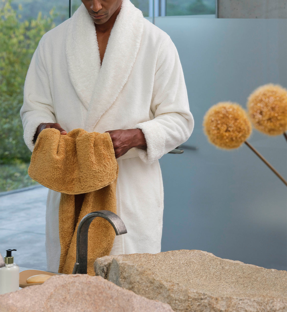 A man in a white bathrobe holds a luxurious premium towel of the finest Egyptian cotton in his hands. The scene radiates calm, elegance and the sensual quality of fine textiles – a symbol of pure relaxation and refined style in the bathroom. Request your free consultation now – Refined bathroom furniture, Tiamont.